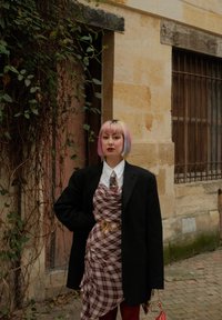 Rutet kjole draperet over en hvid skjorte, lagdelt med en sort oversized blazer. Har et guld kædebelte og levende pink hår.