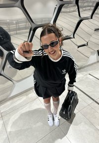 Sweatshirt oversized preto com riscas brancas, combinado com uma saia preta. Acessórios incluem óculos de sol pretos, meias transparentes e uma mala preta.