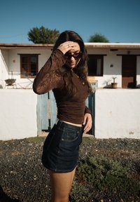 Γυναίκα με γυαλιά ηλίου που διορθώνει τα μαλλιά της, φοράει καφέ μακρυμάνικη μπλούζα και σκουρόχρωμη τζιν φούστα, στέκεται έξω κοντά σε άσπρο κτίριο με μπλε πύλη.