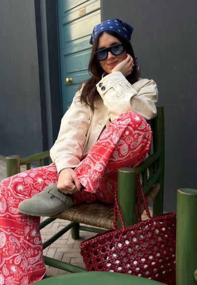 Mujer joven con gafas de sol, pañuelo azul, chaqueta beige y pantalones rojos con estampado, sentada en una silla verde con un bolso tejido rojo cerca.