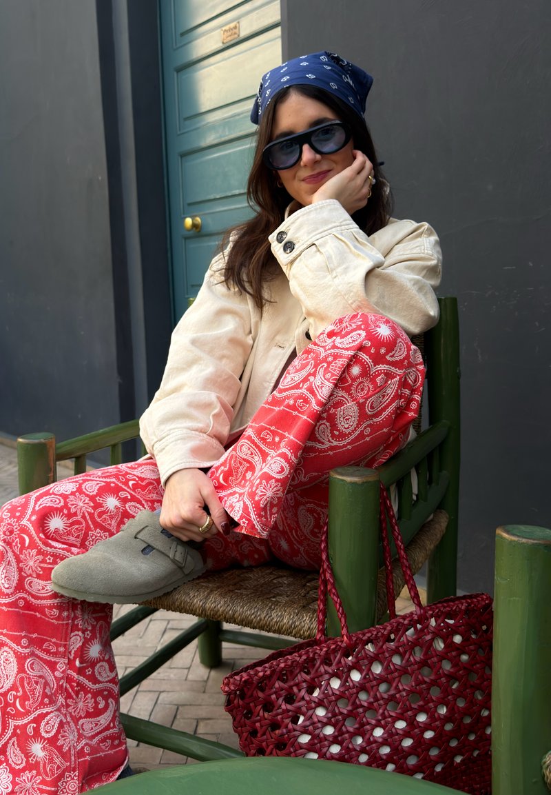 Junge Frau mit Sonnenbrille, blauem Bandana, beiger Jacke und roten gemusterten Hosen, die auf einem grünen Stuhl sitzt, daneben eine rote geflochtene Tasche.