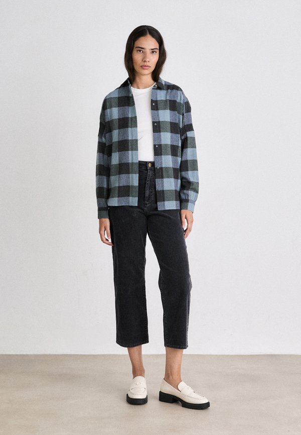 Karierte Bluse in Blau und Schwarz aus Flanell, zugeknöpft, über einem weißen T-Shirt getragen. Schwarze, kurze Hosen und cremefarbene Slipper mit einem niedrigen Blockabsatz.
