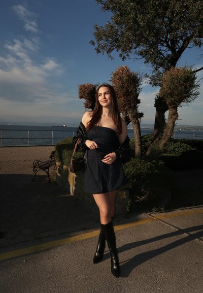 Joven mujer con vestido negro y botas de pie cerca de árboles podados junto a un paseo marítimo bajo un cielo despejado.