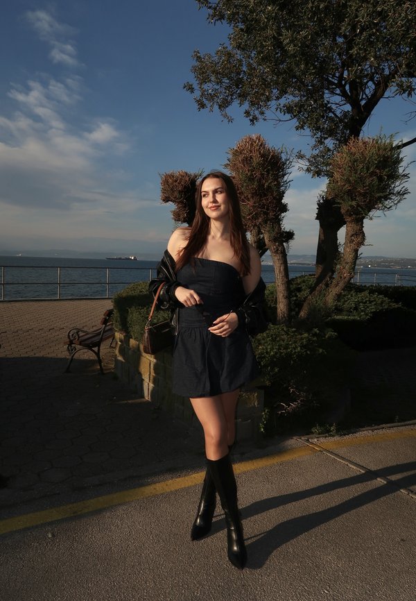 Ung kvinde i sort kjole og støvler står ved trimmede træer ved en strandpromenade under en klar himmel.