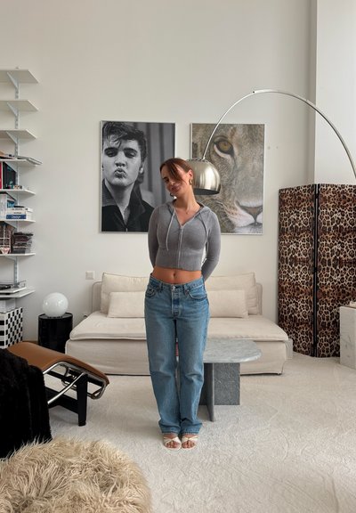 Mujer con sudadera con capucha corta gris y vaqueros, de pie en una sala de estar con sofá beige, dos grandes carteles en la pared, pantalla con estampado de leopardo y lámpara de pie moderna.