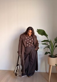 Brauner Trenchcoat über einem gepunkteten Hemd und dunklen, weit geschnittenen Hosen, hält eine gemusterte Handtasche; nahebei eine grüne Pflanze in einem beigen Topf.