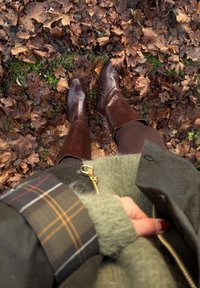 Hnědé kožené kozačky sahající po kolena na posteli z podzimního listí. Zelená bunda s viditelným károvaným vzorem na rukávu.