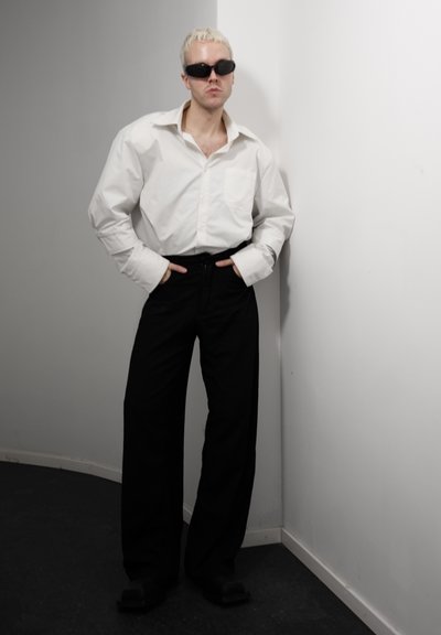 Persona con cabello rubio corto, gafas de sol negras, camisa blanca oversize y pantalones anchos negros apoyada contra una pared blanca.