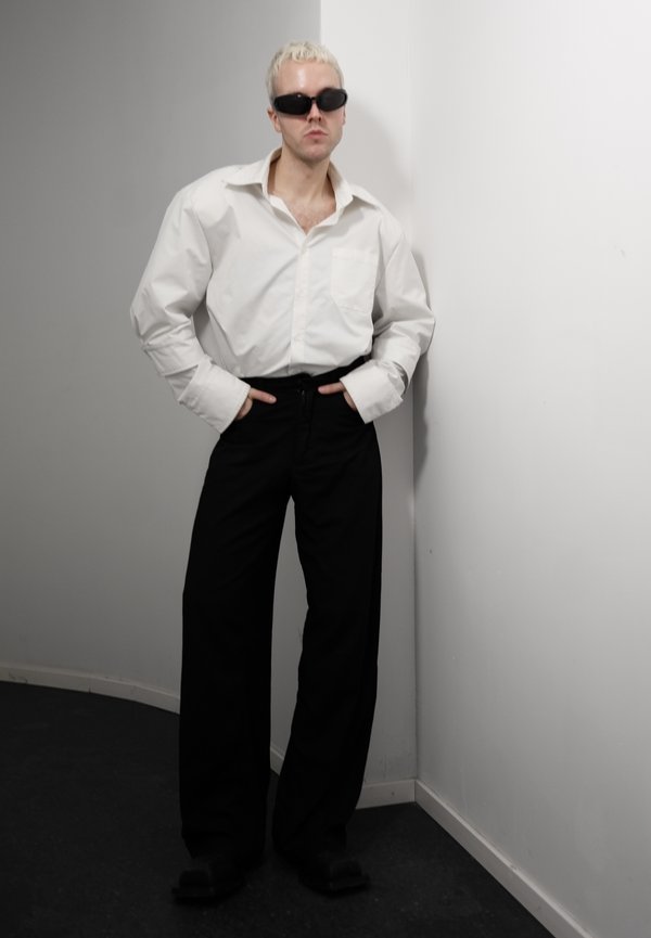 Person mit kurzen blonden Haaren, die schwarze Sonnenbrille, ein übergroßes weißes Hemd und weite schwarze Hosen trägt und sich an eine weiße Wand lehnt.