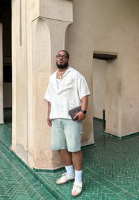 Uformell antrekk med en lys beige kortermet skjorte, denimshorts, hvite sokker og brune joggesko, holder en brun clutch. Tekturert grønn flislagt gulv.