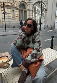 Maglione a motivi argyle verde e marrone, pantaloni in denim a vestibilità rilassata, mocassini in suede marroni e una grande borsa in pelle marrone su un tavolo da caffè.