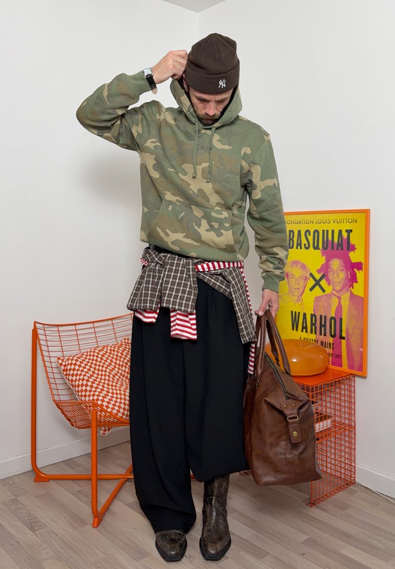Mand iført camouflage hættetrøje, hue, brede sorte bukser, lagdelte skjorter, brune støvler, der holder en lædertaske i et rum med en orange stol og Basquiat-Warhol kunst.