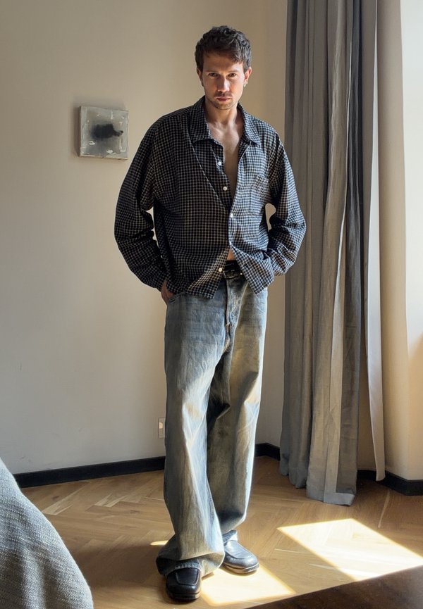 Man standing indoors wearing a loose checkered shirt and wide light-wash jeans with black shoes, near a window with gray curtains.