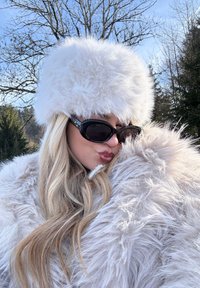 Valge kunstkarusnahast müts ja mantel, millel on pikk, kohev tekstuur; suured mustad p äikeseprillid; välistingimustes, taustal puud ja sinine taevas.