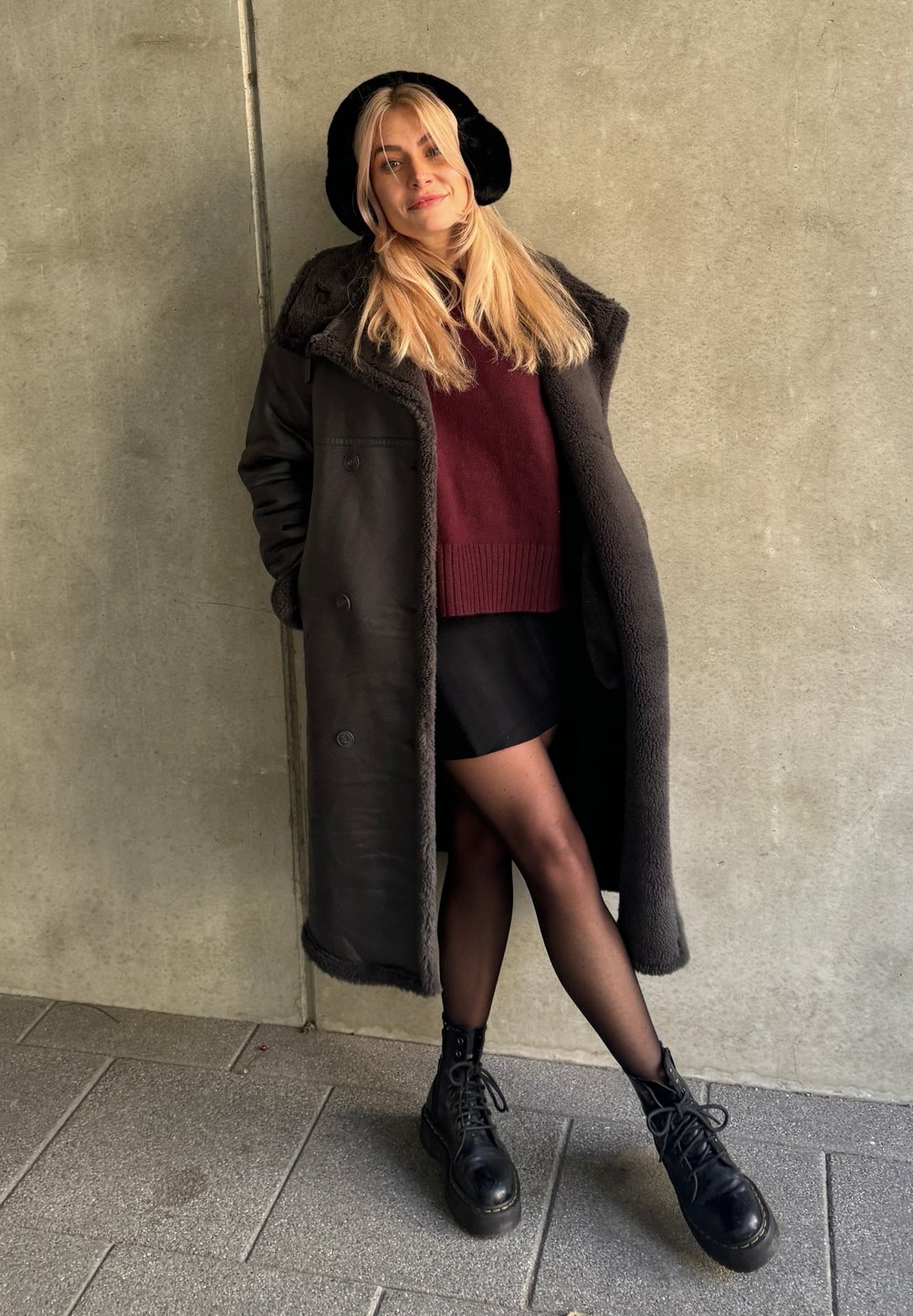 Μαύρο παλτό από μάλλινη ύλη με επένδυση από γούνα, μπορντό πουλόβερ, μαύρη μίνι φούστα και χοντροκομμένες μαύρες μπότες. Στηρίζεται σε έναν τοίχο από σκυρόδεμα.