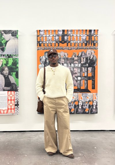 Hombre joven con gafas, gorra marrón, suéter crema y pantalones beige de pie frente a un mural colorido con rostros en blanco y negro.