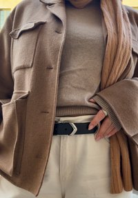 Brauner Wollmantel mit aufgesetzten Taschen über einem beigen Pullover. Ein schwarzer Gürtel mit einer silbernen Schnalle schnürt creamfarbene Hosen. Ein Strickschal aus Kabelmuster ist sichtbar.