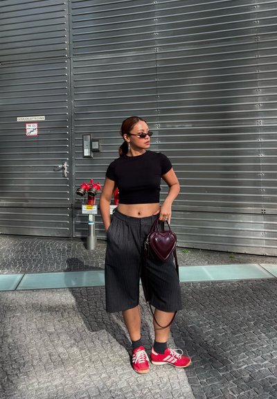 Mujer con top corto negro, culottes de rayas finas, zapatillas rojas, gafas de sol y sosteniendo un bolso en forma de corazón, está junto a una pared de metal con hidrantes rojos.