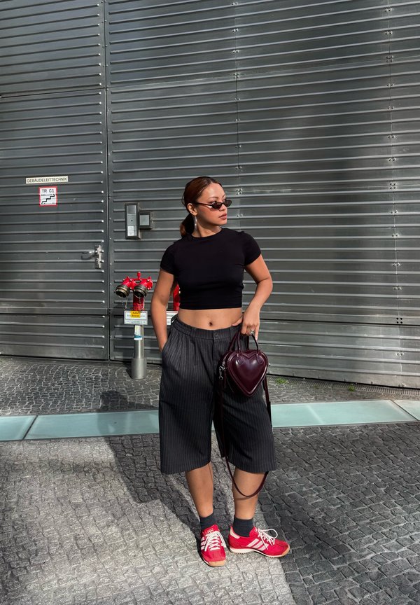 Γυναίκα με μαύρο crop top, culottes με λεπτές ρίγες, κόκκινα αθλητικά παπούτσια, γυαλιά ηλίου και κρατώντας μια τσάντα σε σχήμα καρδιάς στέκεται δίπλα σε μεταλλικό τοίχο με κόκκινες υδρορροές.