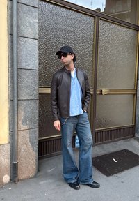 Καφέ δερμάτινο μπουφάν, ανοιχτό μπλε πουκάμισο με κουμπιά, μπλε τζην με φαρδύ σχέδιο, μαύρα παπούτσια, μαύρο καπέλο, γυαλιά ηλίου. Στηρίζεται έναντια σε μια υφασμάτινη φόντο.