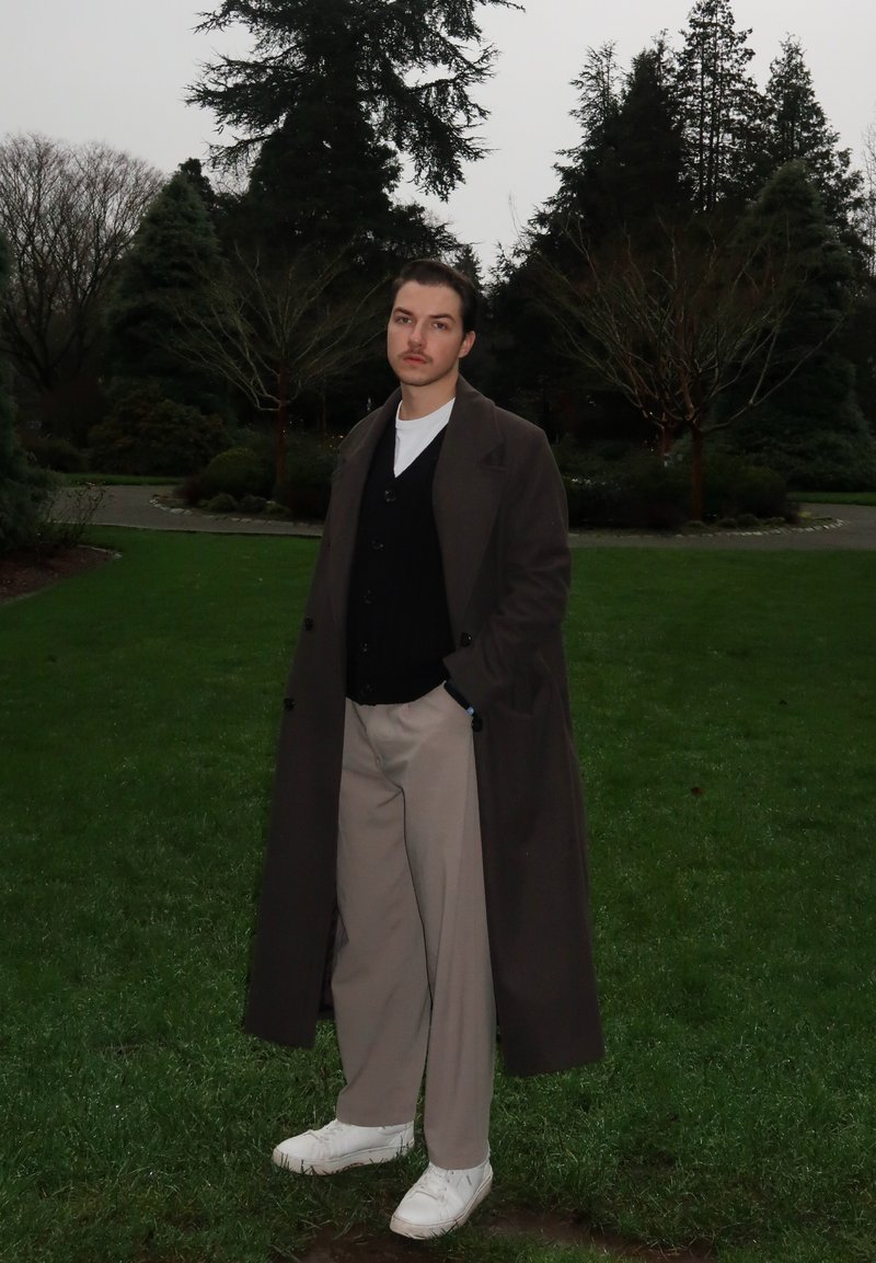 Lange bruine jas, zwarte cardigan, wit overhemd, crèmekleurige broek en witte sneakers. Staand op een grasveld met bomen op de achtergrond.