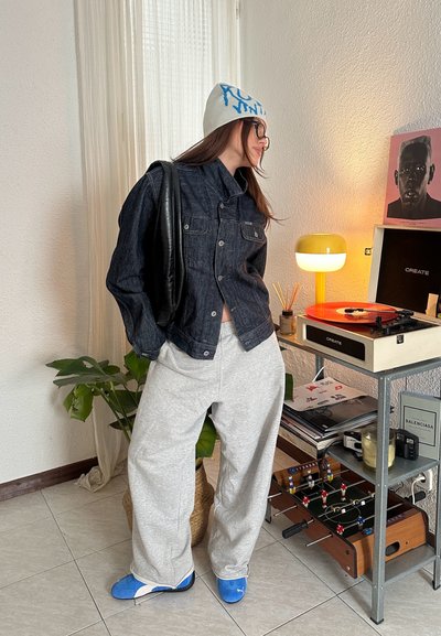 Chaqueta de mezclilla, pantalones de chándal grises, zapatos azules. Los accesorios incluyen un gorro ligero con texto. El fondo presenta un tocadiscos y una planta de interior.