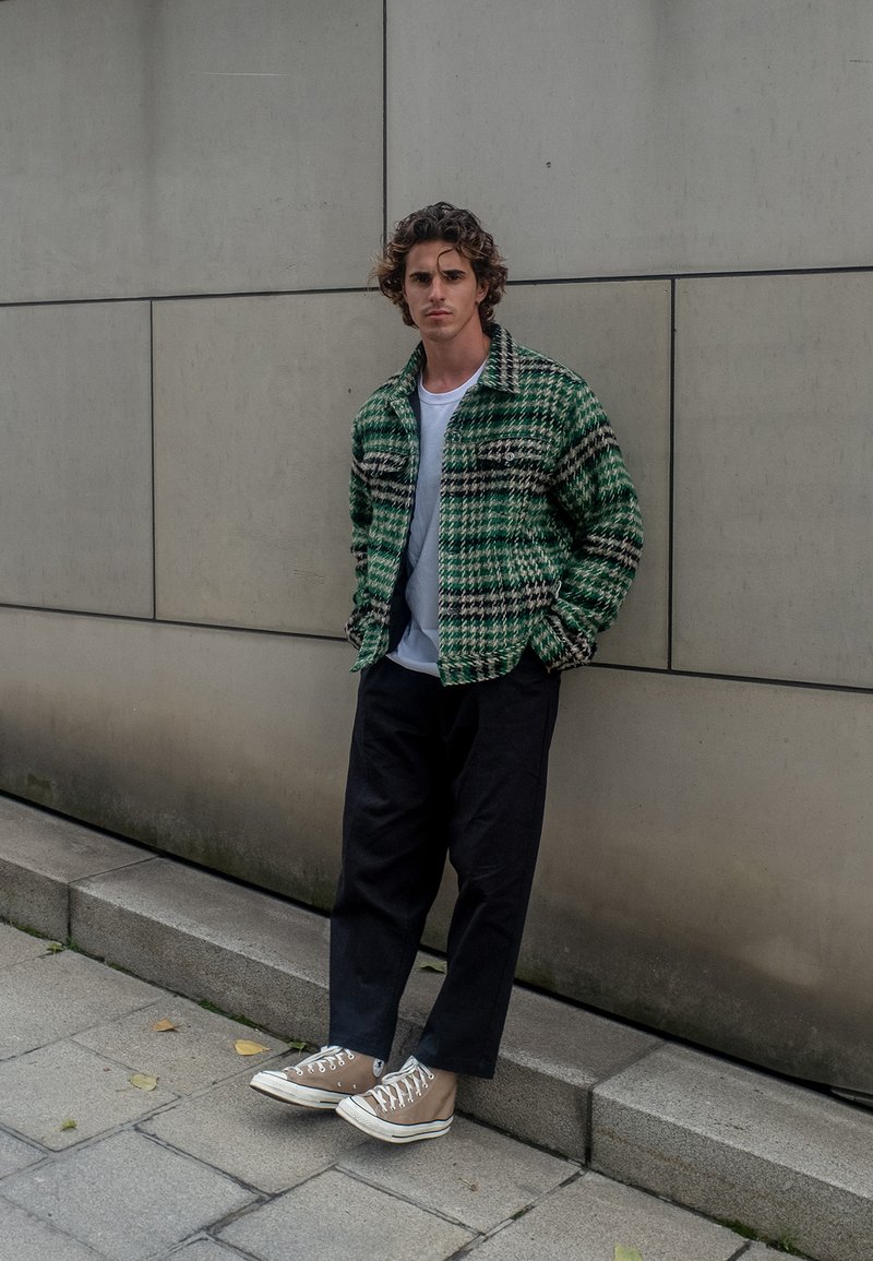 Žalias plaidinė striukė su apykakle, ilgomis rankovėmis, dviem krūtinės kišenėmis, po ja pilkas marškinėliai, juodos kelnės ir smėlio spalvos drobės sportiniai bateliai.