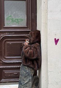 Personne portant une veste en fourrure marron et un pantalon camouflage, buvant un café et s'appuyant contre un mur beige avec une porte marron et un graffiti en forme de cœur à proximité.