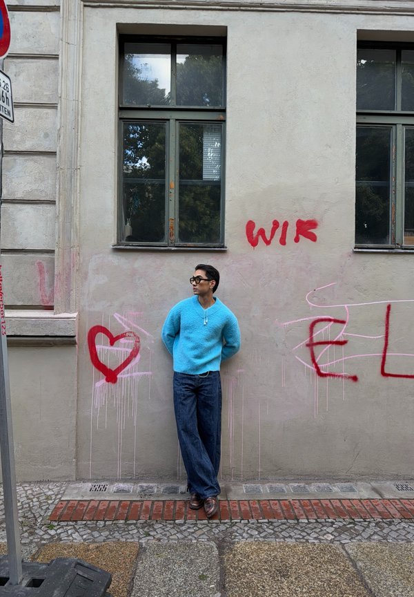 Homme en pull bleu et lunettes de soleil appuyé contre un mur avec un graffiti rouge, incluant un cœur et le mot "WIR."