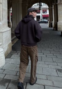 Sweat à capuche oversize marron foncé, pantalon cargo beige avec poches latérales, et chaussures noires. Vue de dos, dans un cadre urbain sous une arche.