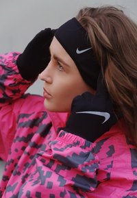 Nike juoda galvos juosta ir pirštinės, rožinė striukė su pilku geometriniu raštu, lengvas audinys, prigludęs dizainas, papuoštas atspindinčiais logotipais.