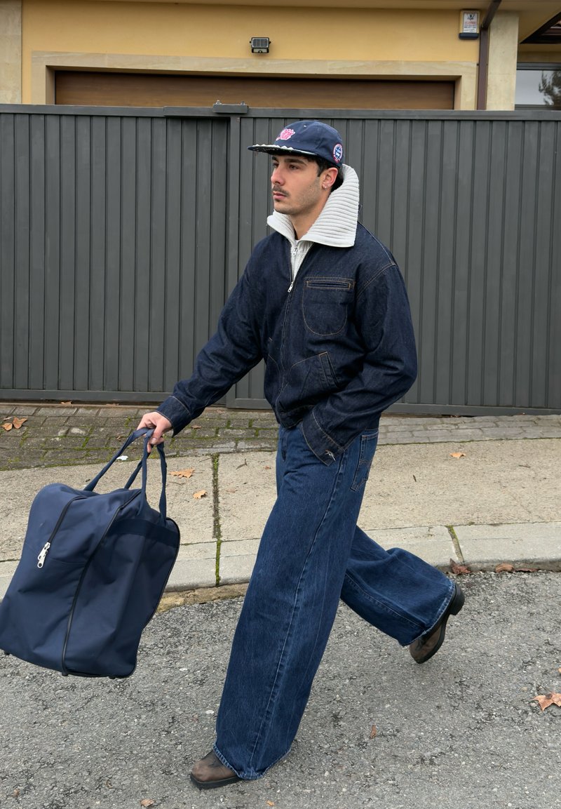 Homme portant une veste en denim foncé et un jean large, marchant et portant un grand sac de sport bleu marine par ses poignées dans une rue pavée.