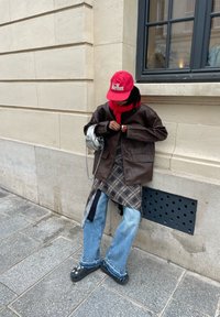 Hnědá kožená oversize bunda, červená čepice, kostkovaná košile, světle modré široké džíny, černé boty se vzorem hvězd, stříbrná crossbody taška.