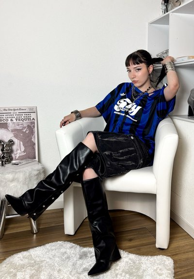 Mujer con cabello negro, vestida con una camisa de rayas azules, falda negra y botas negras hasta la rodilla, sentada en una silla blanca, ajustándose el cabello.