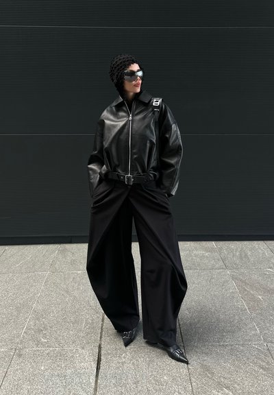 Persona con chaqueta de cuero negra de gran tamaño, pantalones negros de pierna ancha, zapatos puntiagudos, tocado texturizado y grandes gafas de sol reflectantes frente a una pared oscura.