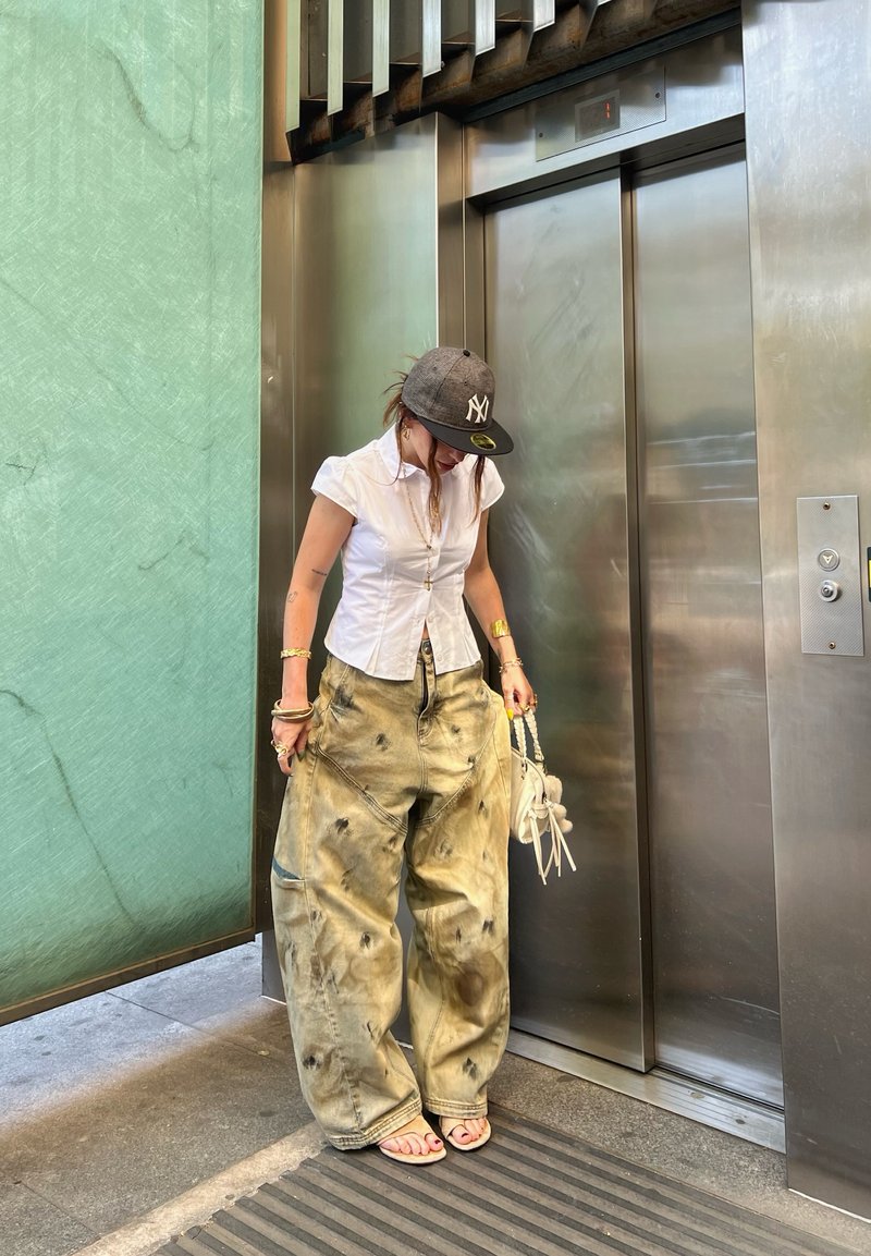 Camisa branca de manga curta com botões, calças largas verdes claras com manchas escuras, boné cinza, pequena mala branca, em pé perto de um elevador.
