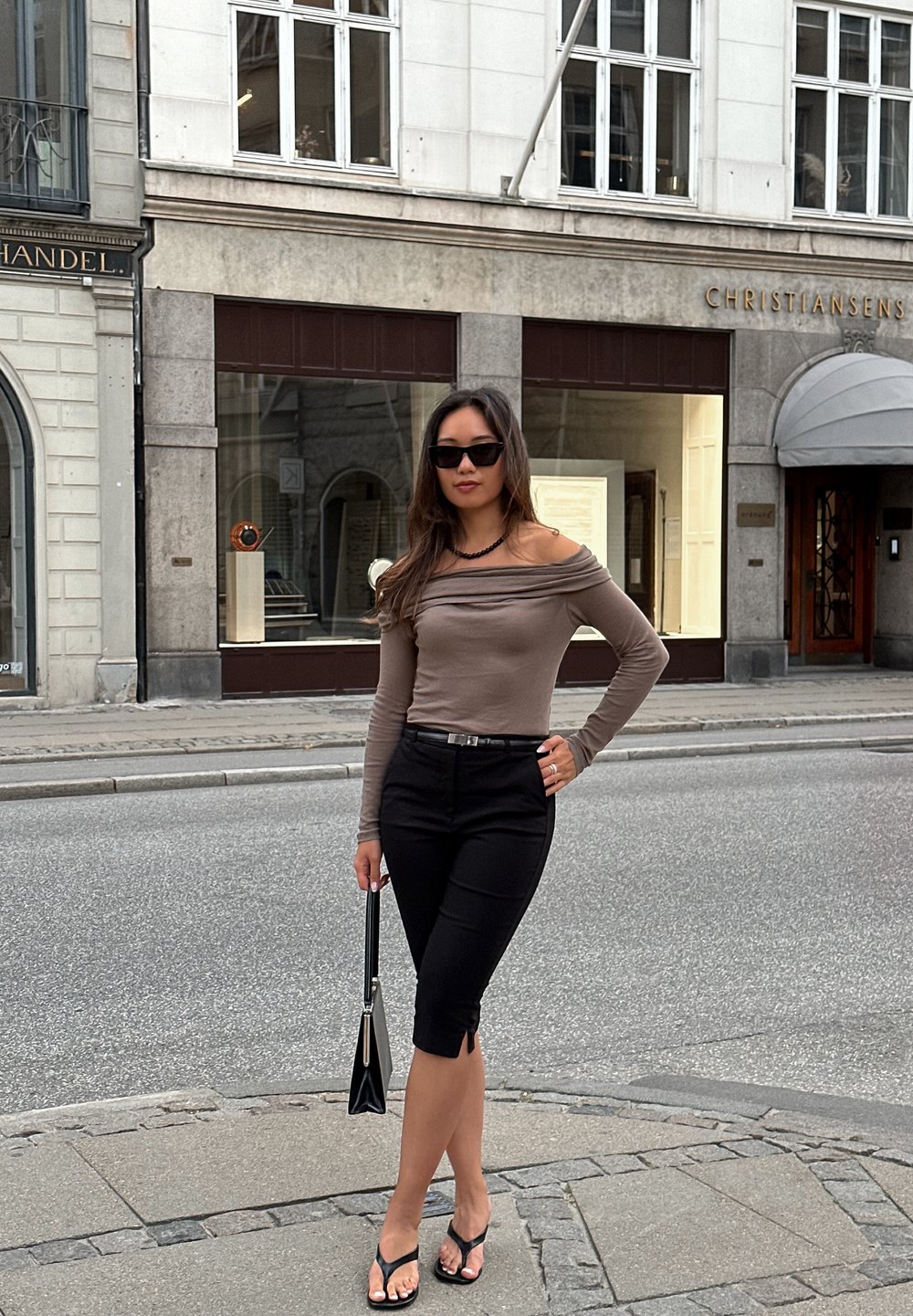 Μακρυμάνικο, εκτός ώμου, ανοιχτό καφέ τοπ, συνδυασμένο με μαύρο κολάν μήκους γονάτου και μαύρες σαγιονάρες. Κρατάει μια μικρή μαύρη τσάντα.