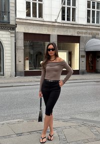 Μακρυμάνικο, εκτός ώμου, ανοιχτό καφέ τοπ, συνδυασμένο με μαύρο κολάν μήκους γονάτου και μαύρες σαγιονάρες. Κρατάει μια μικρή μαύρη τσάντα.