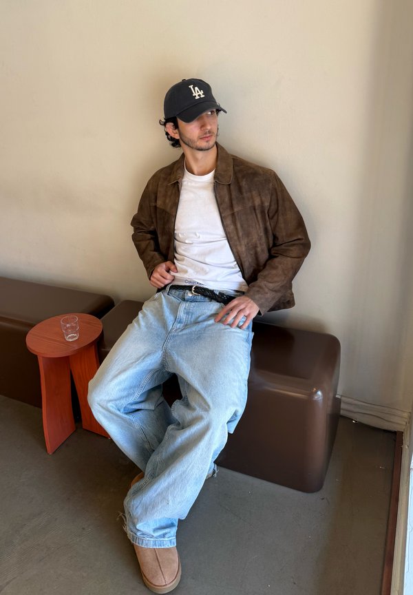 Bruine suède jas, wit T-shirt, verwasemde lichtblauwe jeans en tan kleurige schoenen. Zittend naast een kleine houten tafel met een glas.