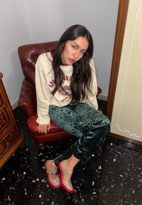Jeune femme aux longs cheveux bruns assise sur un fauteuil en cuir rouge, portant un sweat-shirt crème à motif, un pantalon en velours vert et des chaussures beige à semelles rouges.