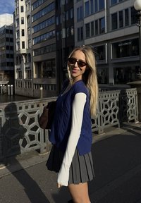 Colete tricotado azul sobre uma camisa de mangas longas branca, combinado com uma saia cinza plissada. Usando óculos de sol escuros, em pé numa ponte.