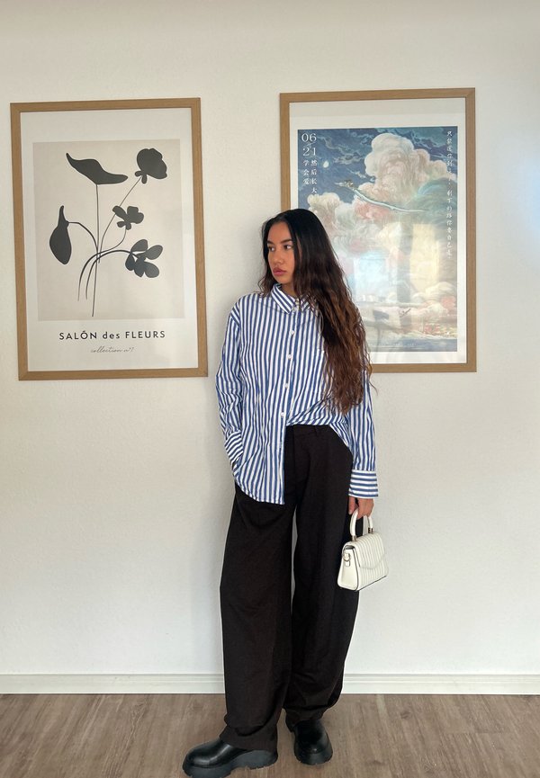 Camisa de botão azul e branca às riscas, calças largas pretas, botas de plataforma pretas, a segurar uma pequena bolsa branca acolchoada. Fundo simples.