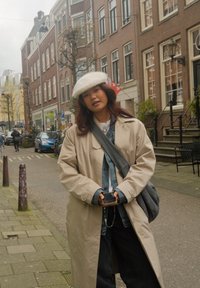 Beige lang frakk, denimskjorte og mørke jeans. Hvit pelslue, grå skulderveske. Bygate med murbygninger og parkerte sykler.