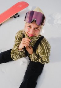 Femme souriante en polaire verte à motifs, pantalon de neige noir et lunettes de ski sur un bonnet écru, assise dans la neige avec un snowboard rouge à côté d'elle.