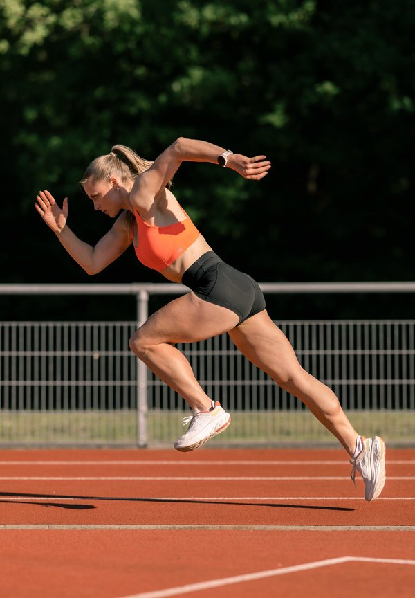 Női sportoló narancssárga sportmelltartóban és fekete rövidnadrágban szalad szabadban lévő futópályán, fél lépésben koncentrált arckifejezéssel, háttérben fákkal.