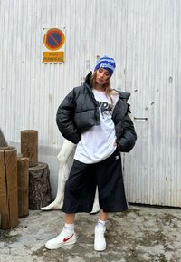 Juodas trumpas pūkinis švarkas, balta grafiška marškinėliai, juodi plačios šortai, balti sportiniai bateliai su raudonais akcentais ir mėlyna megzto skrybėlė su tekstu.