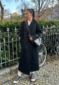 Zwarte dubbelgeknoopte jas, wit overhemd met kraag, zwarte schoudertas, witte kniehoge sokken en zwarte sneakers op een keienstraat.