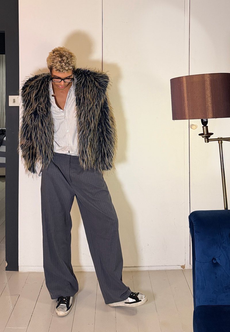 Personne aux cheveux courts et bouclés porte de grosses lunettes, un manteau en fausse fourrure noir et gris duveteux, une chemise à rayures, un pantalon gris ample et des baskets à l'intérieur.