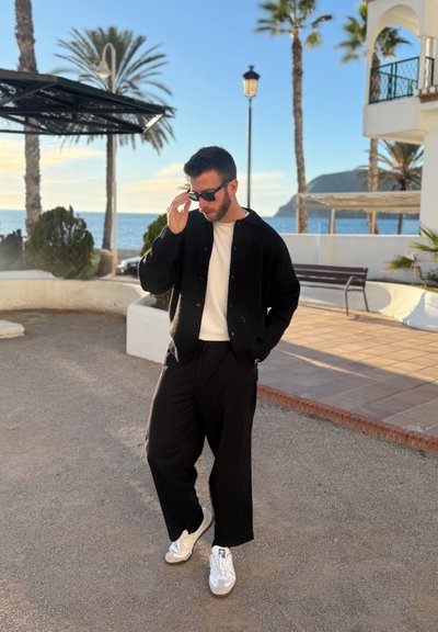 Hombre con cárdigan negro, camisa blanca, pantalones negros y zapatillas blancas ajustándose las gafas de sol cerca de palmeras y el océano al atardecer.