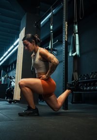 Femme effectuant un fente dans un studio de fitness. Elle porte un crop top beige et un short orange, avec des chaussures de sport noires. Équipement de gym en arrière-plan.